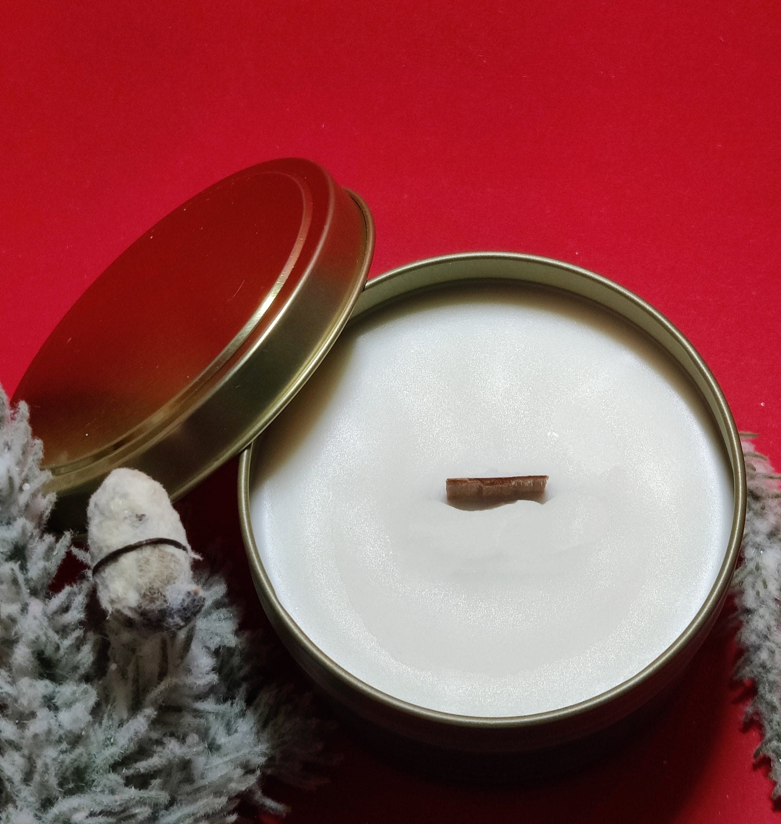 Open gold tin candle with a brown wick on a white wax base, surrounded by winter decor on a red background.