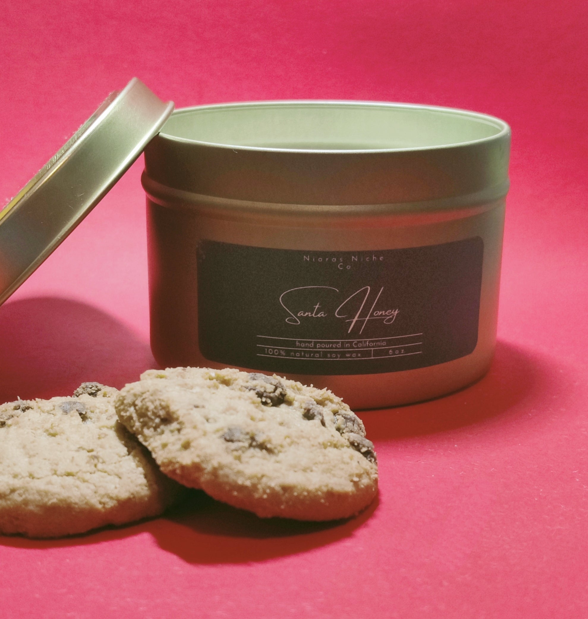 Santa Honey scented candle in a metal tin, hand poured in California, with two chocolate chip cookies nearby.